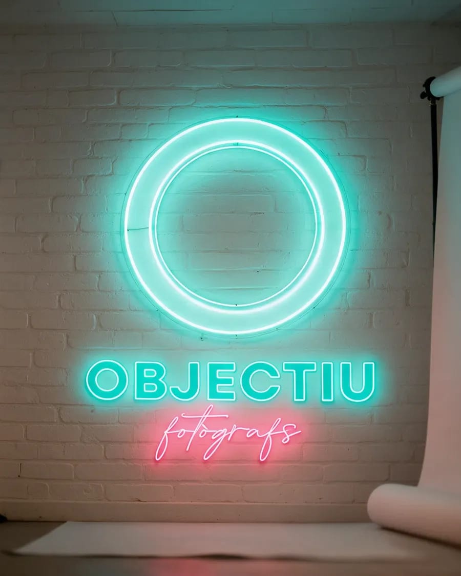 Logotip d'Objectiu Fotògrafs en neó sobre una paret de maó, entre dos focus d'estudi